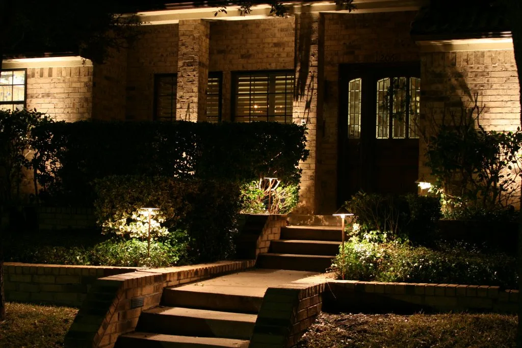 Warm light illuminates a welcoming brick house and its landscaped surroundings at night