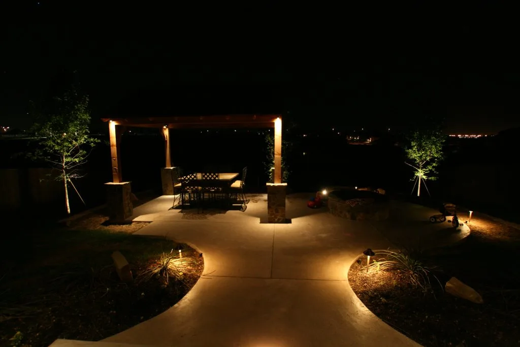A softly lit patio features a wooden pergola, surrounded by gentle landscaping at night