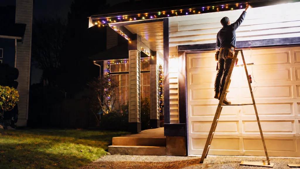 A person stands on a ladder, decorating a house with holiday lights at night